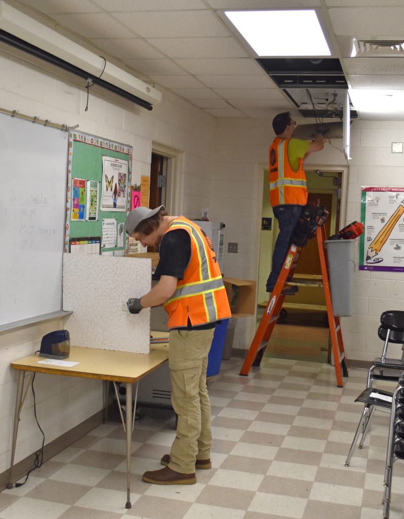 Two men work to replace ceiling lights in a classroom