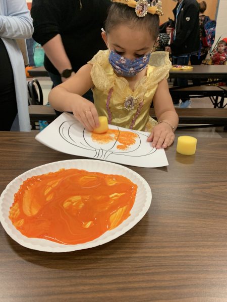 A young girl uses a sponge and a plate of orange paint to color a pumpkin picture during a 2021 Parent University class.
