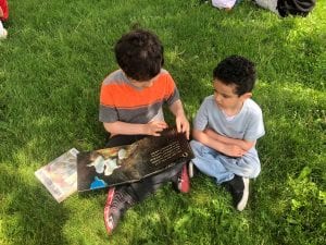 A fourth grade boy reads to a preschool boy outside, sitting on the grass.