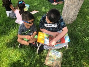A fourth grade boy reads to a preschool boy outside, sitting on the grass.