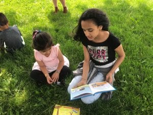 A fourth grade girl reads to a preschool girl outside, sitting on the grass.