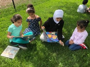 Two fourth grade girls read to two preschool girls outside, sitting on the grass.