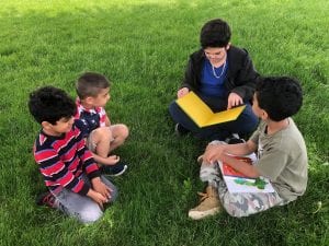 Two fourth grade boys read to two preschool boys outside, sitting on the grass.
