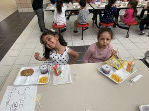 Two preschool girls smile while eating their lunch in the cafeteria.