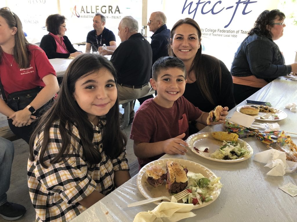 Mrs. Younes, Ali, and Ayat enjoy burgers and corn on the cob while smiling at the camera.