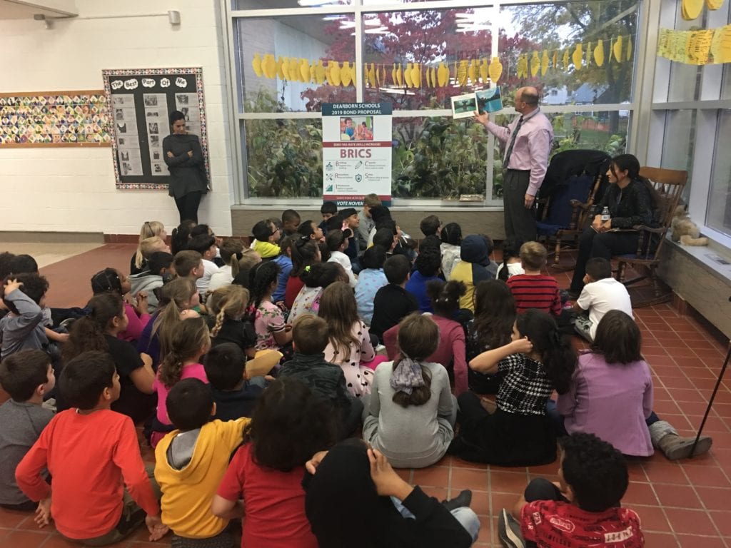    School Board Member Jim Thorpe reads a Halloween story to 2nd graders at Snow! He is standing holding a book up, while the principal sits in a rocker next to  him. Students are seated on the floor in front of  him. A teacher is standing to the front left of the reader with her arms crossed , while she looks down. 