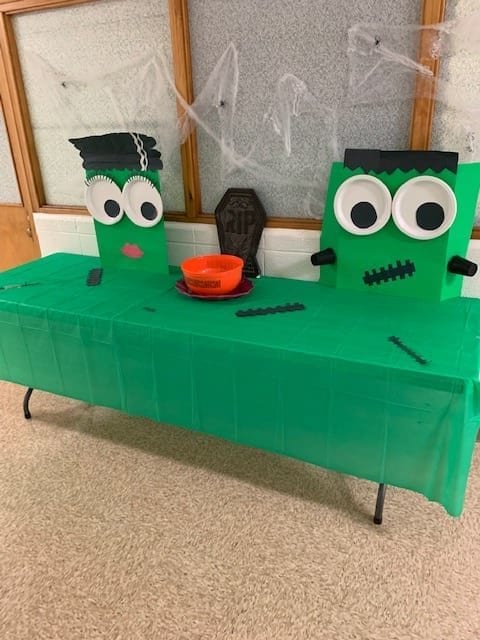 Halloween theme decorated cafeteria table all decorated in a green Frankenstein theme in the hallway. There are spider webs hung behind the table, and an orange candy bowl as well a pretend headstone on the table.