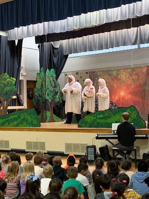 Three adult actors, all dressed in white goat costumes appear to be talking on stage. Students are sitting on the gym floor, watching the play. 