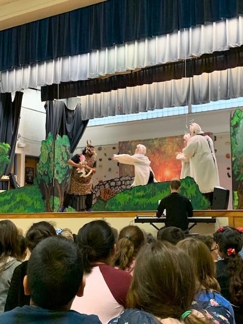 Four adult actors are on stage. From left is the troll, clutching the goat's stuffed rabbit, then the three billy goats. The nearest goat has  her arms outstretched trying to get her rabbit back from the troll. Snow students are seated on the floor of the gym. 