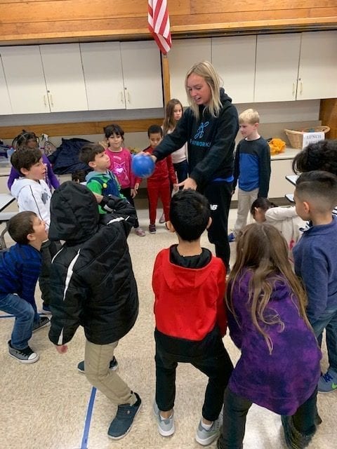 Female PlayWorks coach wearing a dark PlayWorks hoodie, holding a blue ball, surrounded by a circle of children. She holds the ball out to a student.