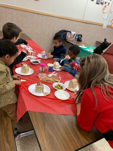 Many children sit at cafeteria tables covered in red or green tablecloths. Each child has a plate  with a small gingerbread house on it in front of him or  her, along with many bowls of toppings for the children to attach to the gingerbread houses with white frosting. Mostly the gingerbread houses look bare so far as they are just getting started on this project. Teachers and parents are spread between the tables of children to assist and guide the project. 