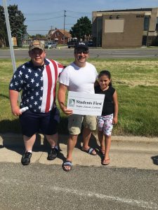 2 adults and a student with the sign at the parade