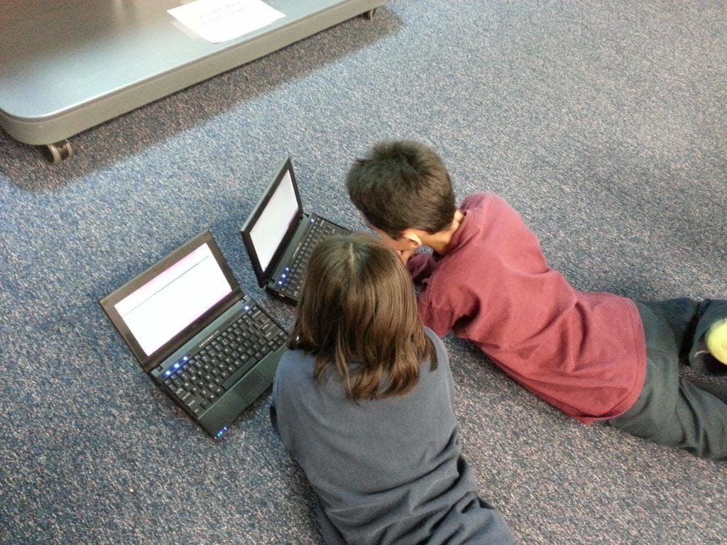 two students, a boy and a girl, lay on the carpeted floor and work on chromebooks
