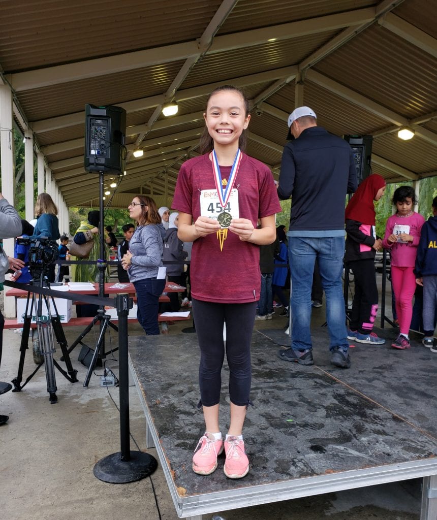 Calyssa Standing on a platform wearing a medal around her neck while holding the medal with both hands.  She has on a maroon shirt, black pants and pink shoes.  Her hair is pulled back into a pony tail.