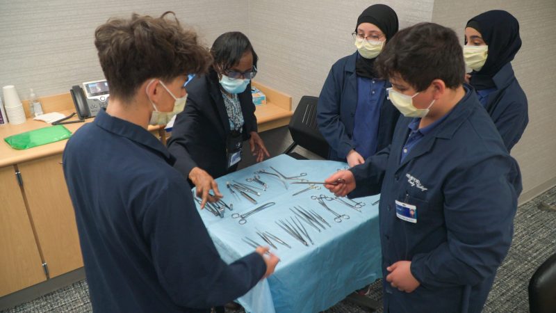 Four Henry Ford Early College students and a woman stand around a tray of surgical instruments as she teaches them about the tools and how to handle them.