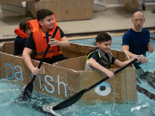 STEM students paddle their cardboard boat across a pool in 2024