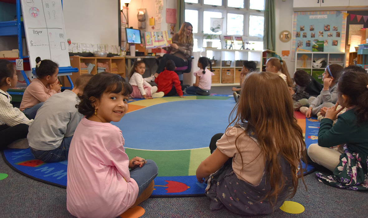 Students sit in a circle listening to a teacher read a story.