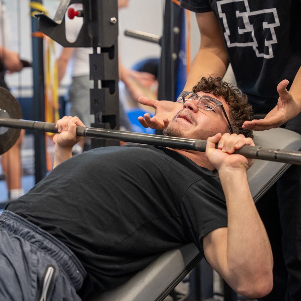 Student weightlifting on bench press with student standing behind and supporting.